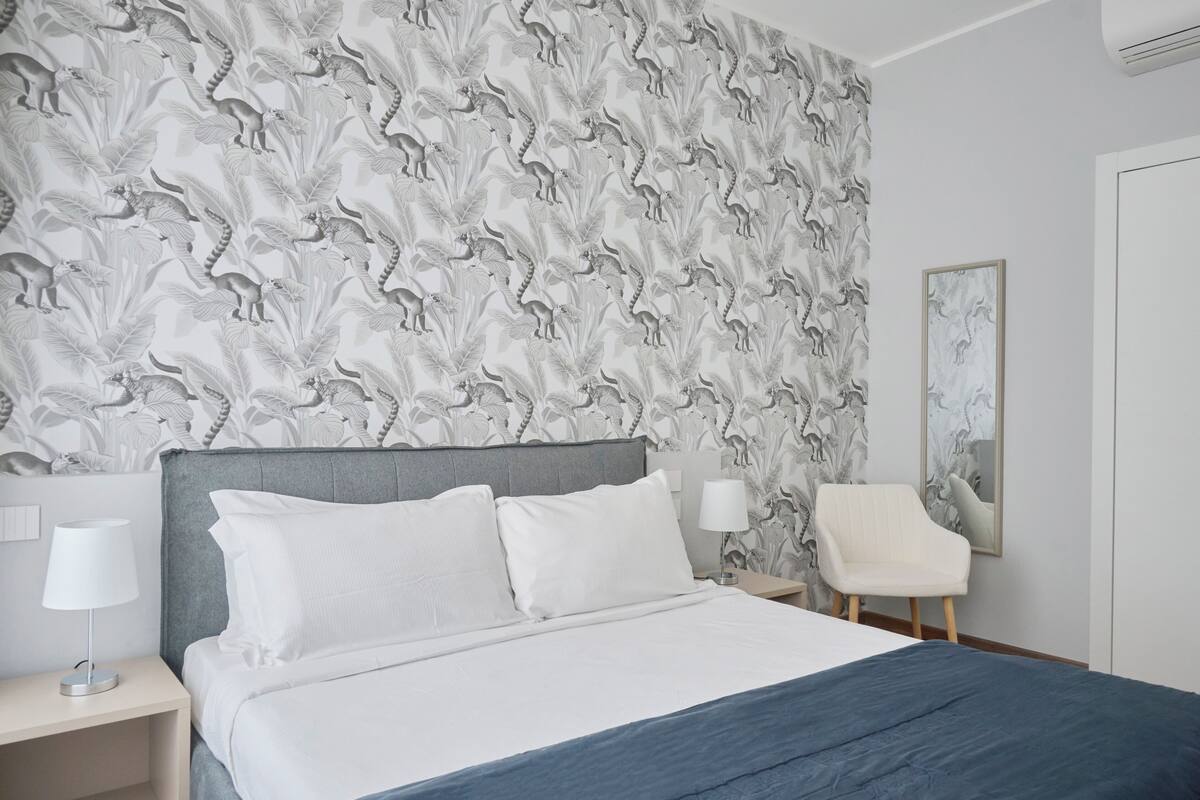 Habitación de hotel con papel tapiz estampado, una cama con ropa de cama blanca, un cabecero gris, una mesita de noche con lámpara y una silla.