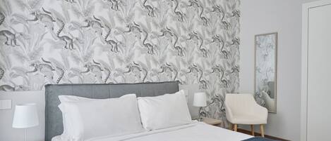 Habitación de hotel con papel tapiz estampado, una cama con ropa de cama blanca, un cabecero gris, una mesita de noche con lámpara y una silla.