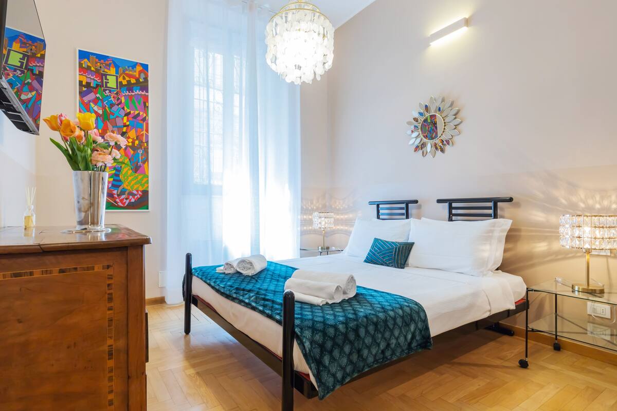 Un dormitorio moderno con cama, mesitas de noche, una araña y obras de arte en las paredes.
