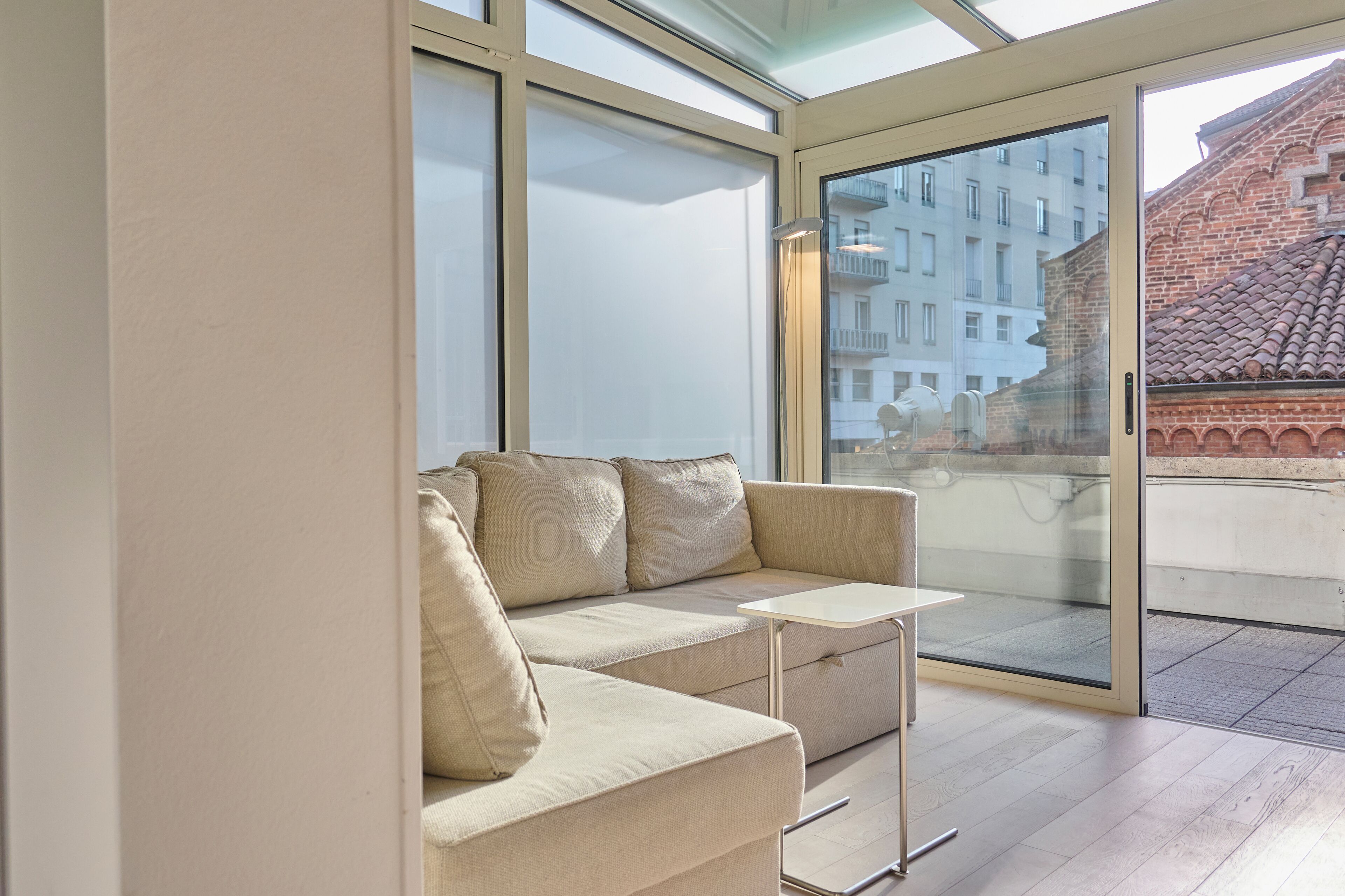 A modern living room with a beige sofa, a small white table, and a large sliding glass door leading to an outdoor balcony.