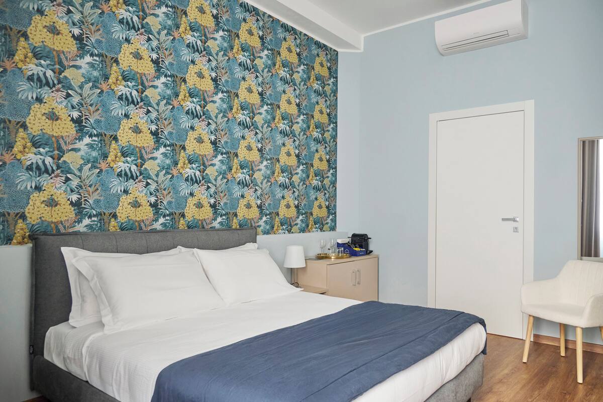 Un dormitorio con una cama, una mesita de noche, una silla, un armario y un papel tapiz con motivos florales.