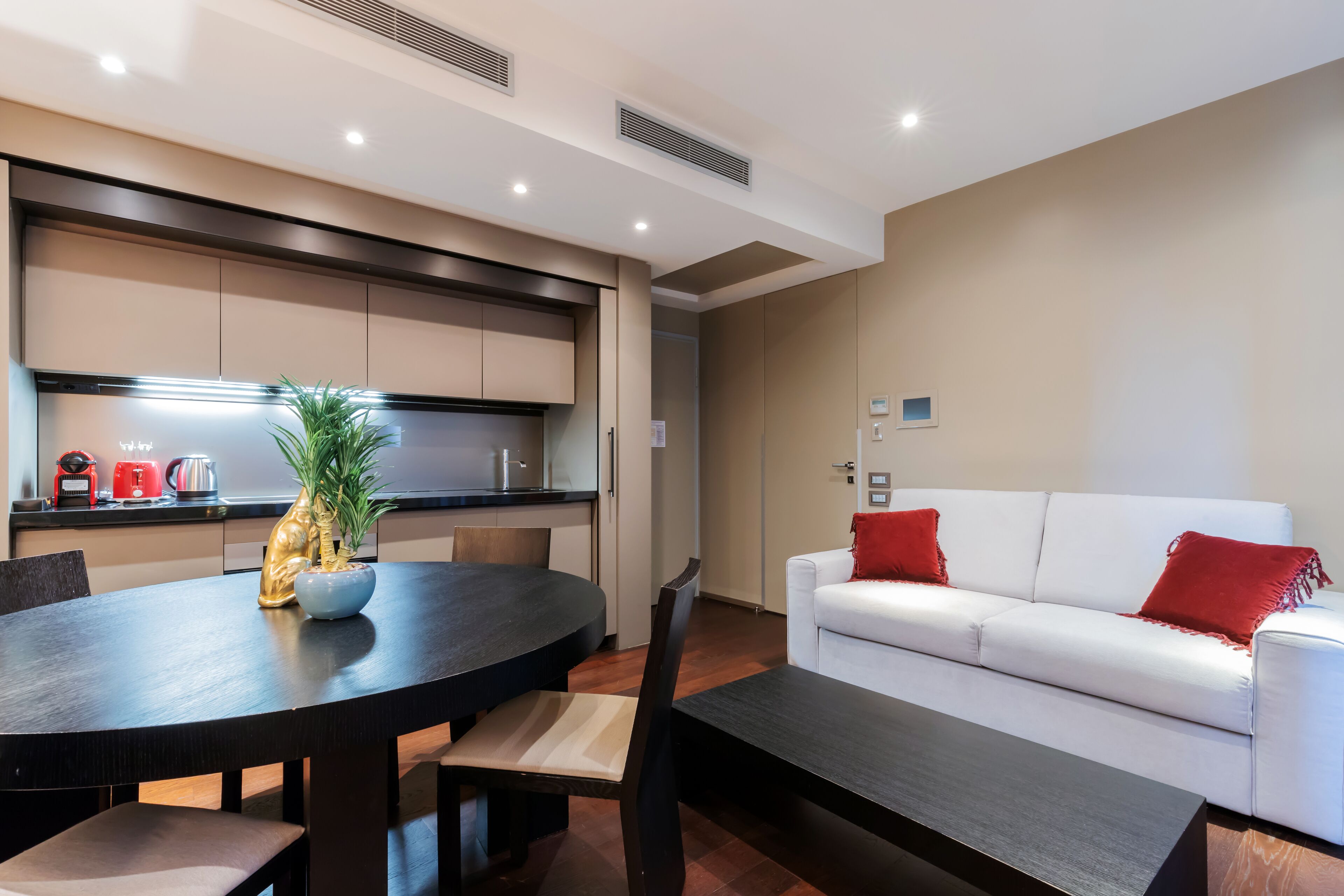 A modern living room with a white sofa, red cushions, a dining table with chairs, and a kitchen area with cabinets and a plant.