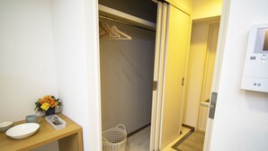 A hotel room with a wooden desk, a white bowl, a cup, a vase with flowers, a laundry basket, and a closet with hanging clothes. - Grande Shagi GS206 (Osaka)