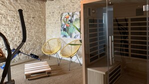 A room with a stone wall, wooden ceiling beams, a massage table, two wicker chairs, and a glass-enclosed sauna. - La Belle Tuilière: A Charming Home in Champagne.  (Vandières)