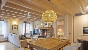 Een gezellige woonkamer met een open haard, houten balken in het plafond, een eettafel met stoelen en een groot raam met gordijnen.