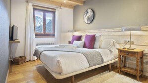 Een slaapkamer met een bed, nachtkastje, lamp, klok en een raam met uitzicht op bergen.