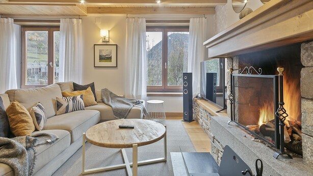 Een gezellige woonkamer met een open haard, een bank, een bijzettafeltje en uitzicht op de natuur door grote ramen.