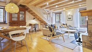 Een ruime woonkamer met een houten trap, een eettafel met stoelen, een bank en een groot raam.