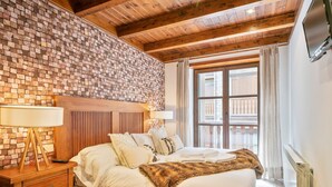 A bedroom with a brick-patterned wall, wooden ceiling beams, a large window with curtains, a bed with white linens, and a nightstand with a lamp. - Luderna - Apartamento Val de Ruda B1 Morèdo (Baqueira)