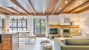 A spacious living area with a fireplace, wooden ceiling beams, and a large window overlooking greenery. - Luderna - Apartamento Val de Ruda B1 Morèdo (Baqueira)