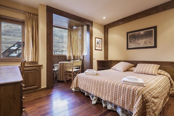 A bedroom with a large bed, a desk, and a chair. There is a window with curtains, a painting on the wall, and a view of a building outside.