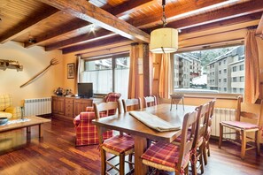 A dining area with a wooden table and chairs, a hanging lamp, and a view of buildings through large windows. - Luderna - Apartamento Tuca (Vielha)