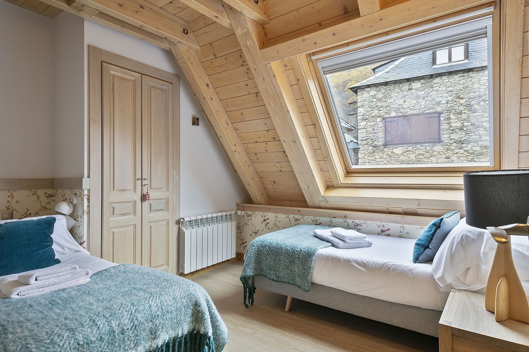 Slaapkamer met schuine plafond, een stenen muur en een raam met uitzicht op een stenen gebouw. Er staan twee bedden met blauw-witte beddengoed, een houten bankje en een radiator.