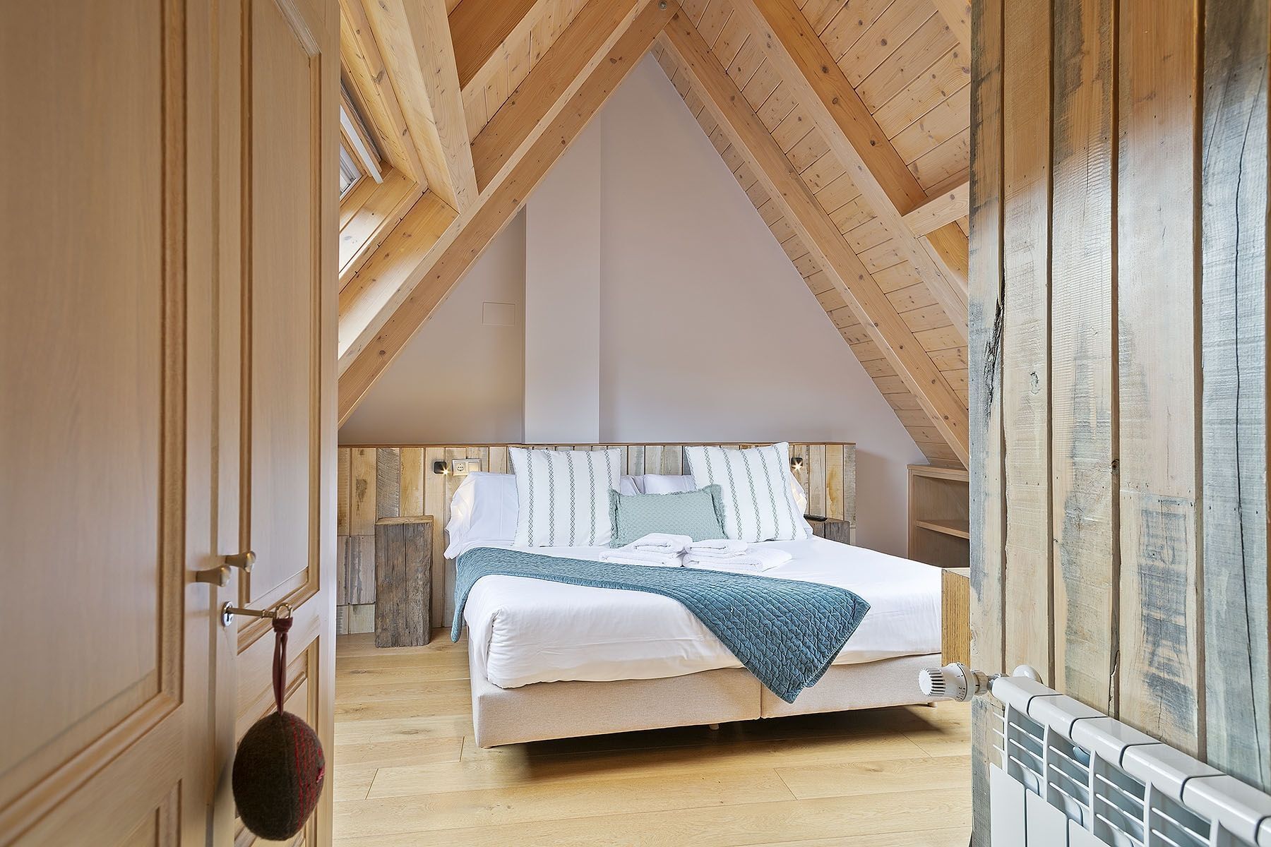 A bedroom with a high wooden ceiling, a bed with white linens and a blue blanket, and a radiator on the right.