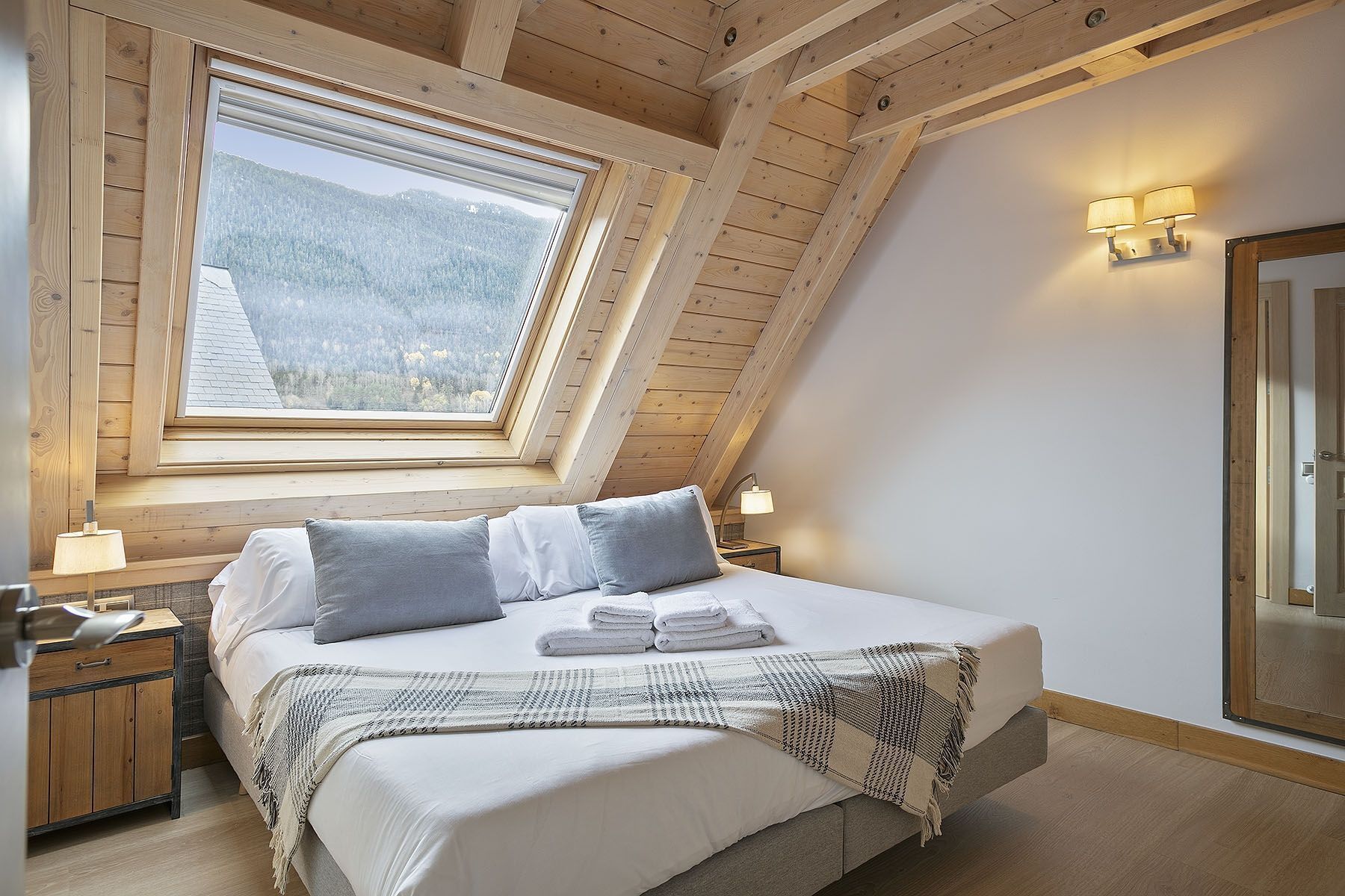 Une chambre avec un plafond en pente, une grande fenêtre donnant sur une montagne boisée, un lit avec des oreillers et une couverture, des lampes de chevet et une table de chevet en bois.