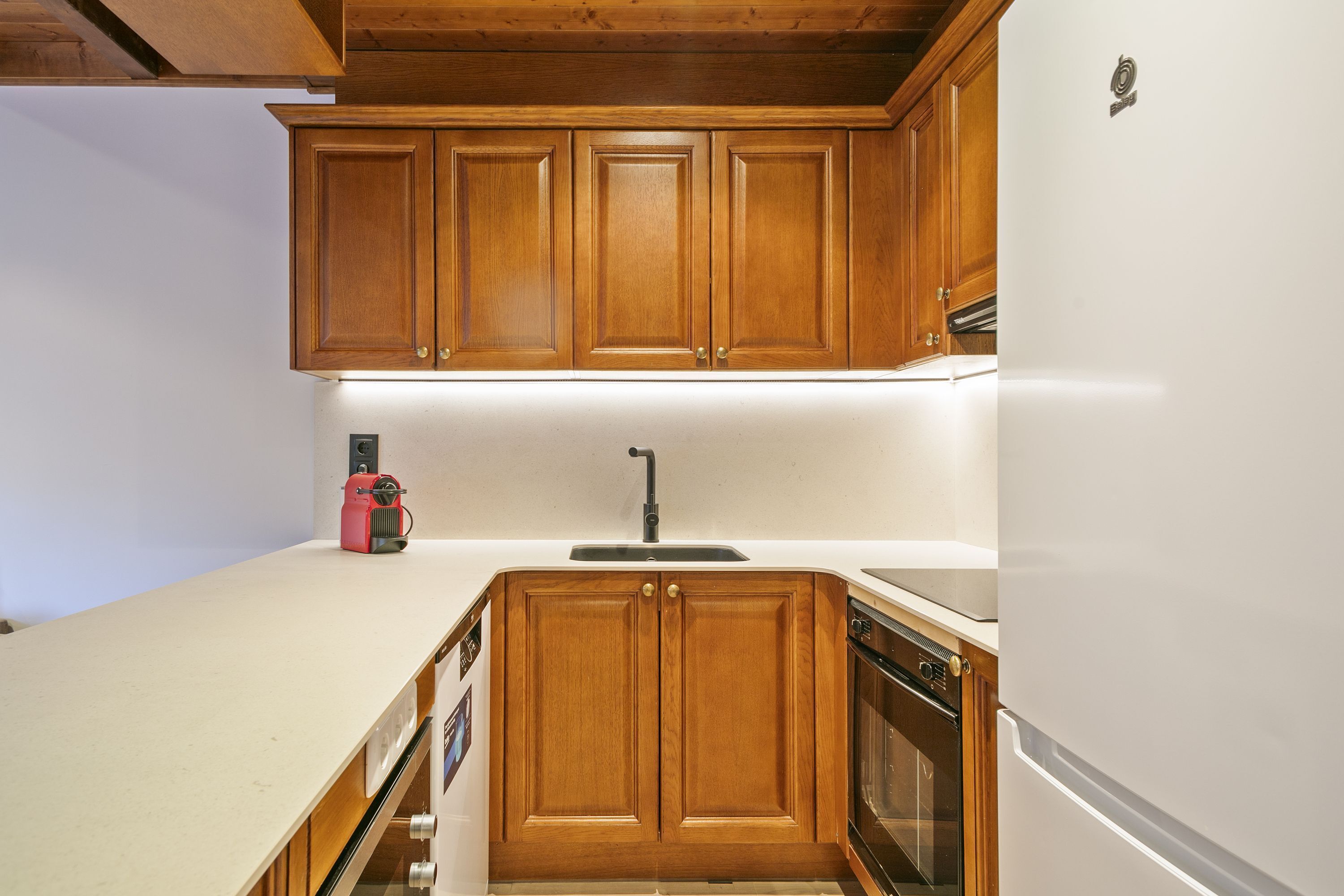 Una cocina con armarios de madera, una encimera blanca, un fregadero y un refrigerador.