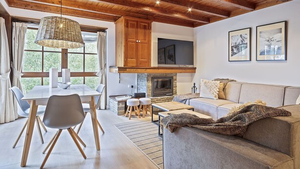 Un salon moderne doté d’une cheminée, de poutres apparentes en bois au plafond, d’un coin repas avec une table et des chaises, et d’un canapé agrémenté d’un plaid.