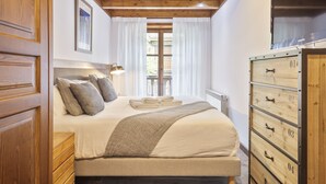 A bedroom with a wooden bed, a bedside table, a lamp, a window with curtains, and a wardrobe with numbered drawers. - Luderna - Apartamento Val de Ruda A6 Porèra (Baqueira)