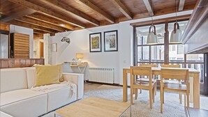 A cozy room with a wooden ceiling, a white sofa with a yellow throw, a wooden coffee table, and a dining area with wooden chairs and a table. - Luderna - Apartamento Val de Ruda A6 Porèra (Baqueira)