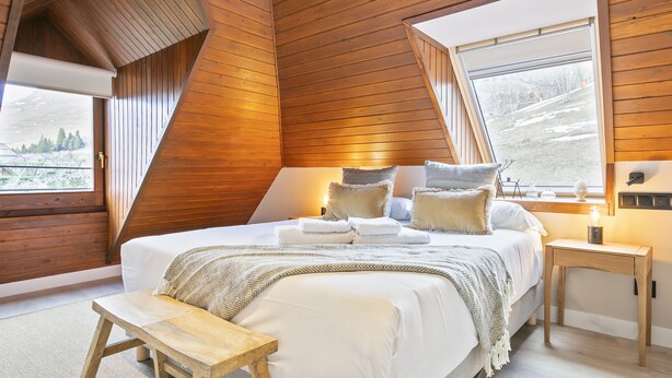 A bedroom with a sloped ceiling, a large bed with pillows and a blanket, a bedside table with a lamp, and a window with a view of the outdoors.