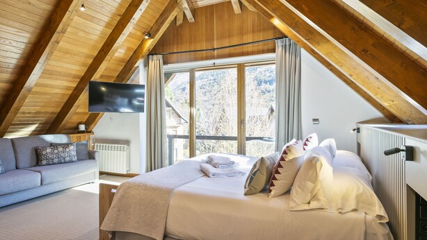 Une chambre avec un plafond en bois, un lit avec des oreillers et une couverture, un canapé avec des coussins, un téléviseur fixé au mur et une fenêtre donnant sur les arbres.