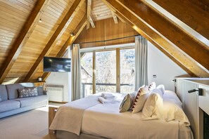 Een slaapkamer met een houten plafond, een bed met kussens en een deken, een bank met kussens, een televisie aan de muur en een raam met uitzicht op bomen.