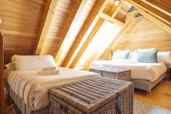Un dormitorio cálido en el ático con techo de madera, dos camas, mobiliario de mimbre y un ventanal grande.