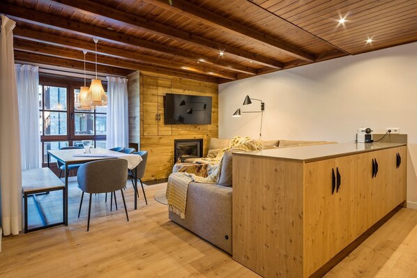 Quarto acolhedor e bem iluminado com teto de madeira, área de jantar com mesa e cadeiras, sofá e televisão fixada na parede.