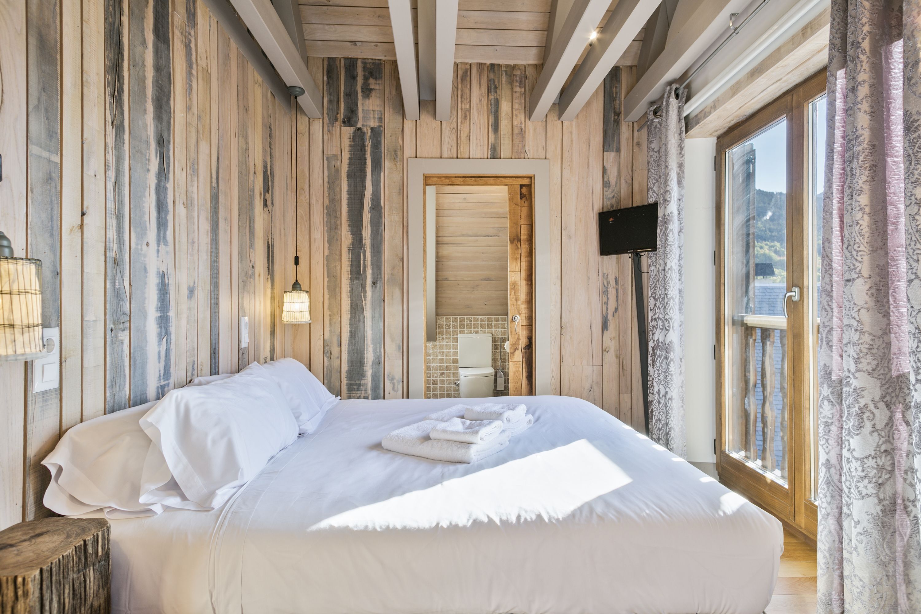 Une chambre avec un mur en bois, un lit recouvert de draps blancs, un téléviseur fixé au mur et une fenêtre avec des rideaux.