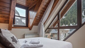 Ein Schlafzimmer mit schrägen Wänden, Holzbalken, einem großen Fenster mit Blick auf Bäume, einem Bett mit weißen Leinen und einer Heizung.