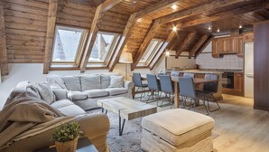 Ein Loft-Wohnbereich mit Holzbalkendecke, einer Couch, einem Esstisch mit Stühlen und einer Küche im Hintergrund.