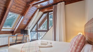 Ein Schlafzimmer mit einem Bett, einem Schreibtisch und einem Stuhl. Fenster mit Vorhängen bieten Blick auf Bäume.