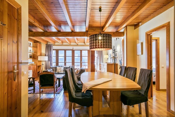 Un comedor de madera con una mesa grande y sillas, una lámpara colgante y vistas al exterior a través de amplias ventanas.