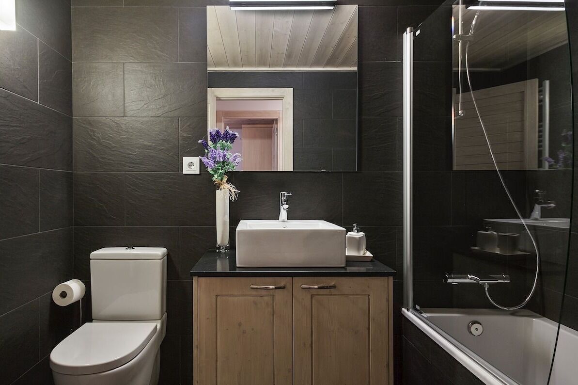 Un bagno moderno con piastrelle scure, un water bianco, un mobile lavabo in legno con specchio e un'area doccia con pareti in vetro.