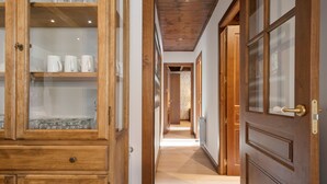 A wooden hallway with glass-fronted cabinets, a door with a brass handle, and a patterned wall in the background. - Luderna - Apartamento Val de Ruda B21 Blanhiblar (Baqueira)
