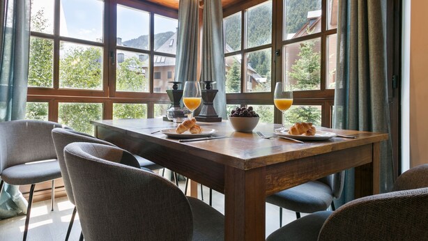 A wooden dining table with chairs, a bowl of fruit, croissants, and glasses of orange juice. - Luderna - Apartamento Val de Ruda B21 Blanhiblar (Baqueira)