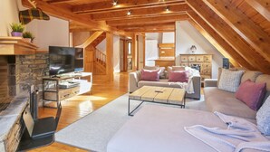 Ein gemütliches Loftsesselzimmer mit Kamin aus Stein, Holzbalken, einer Couch und einem Fernseher.