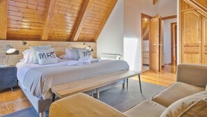 Ein Schlafzimmer mit Holzbalkendecke, einem Bett mit Kissen mit der Aufschrift 'FRAU' und 'HERR', einem Nachttisch, einem Sessel und einer Bank.