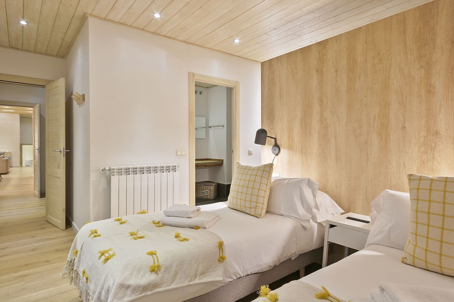 A bedroom with a wooden ceiling, a white radiator, a bedside table with a lamp, and a bed with white bedding and yellow accents.