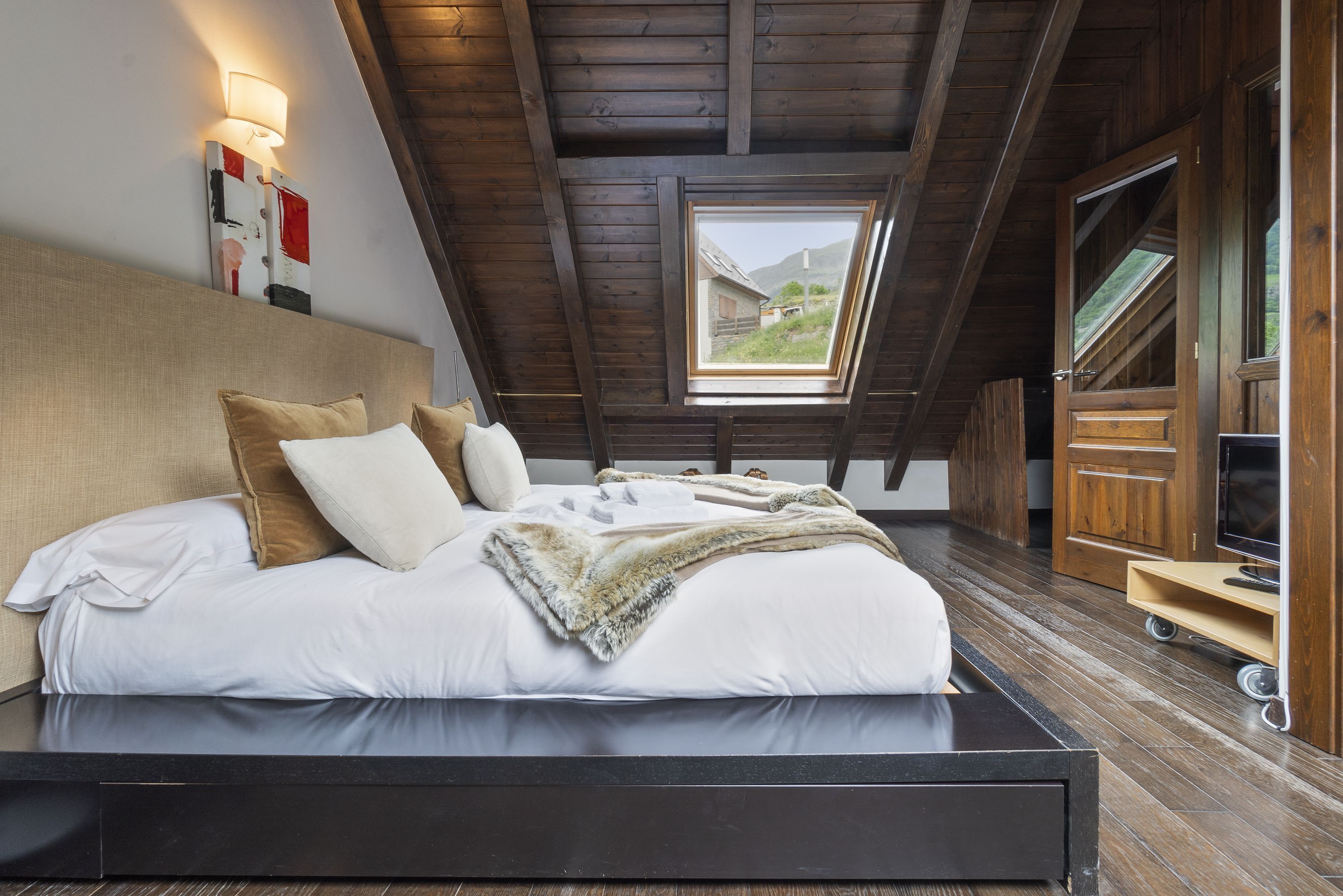 Un dormitorio con techo de madera, un tragaluz, una cama con ropa de cama blanca y un televisor sobre un mueble de madera.