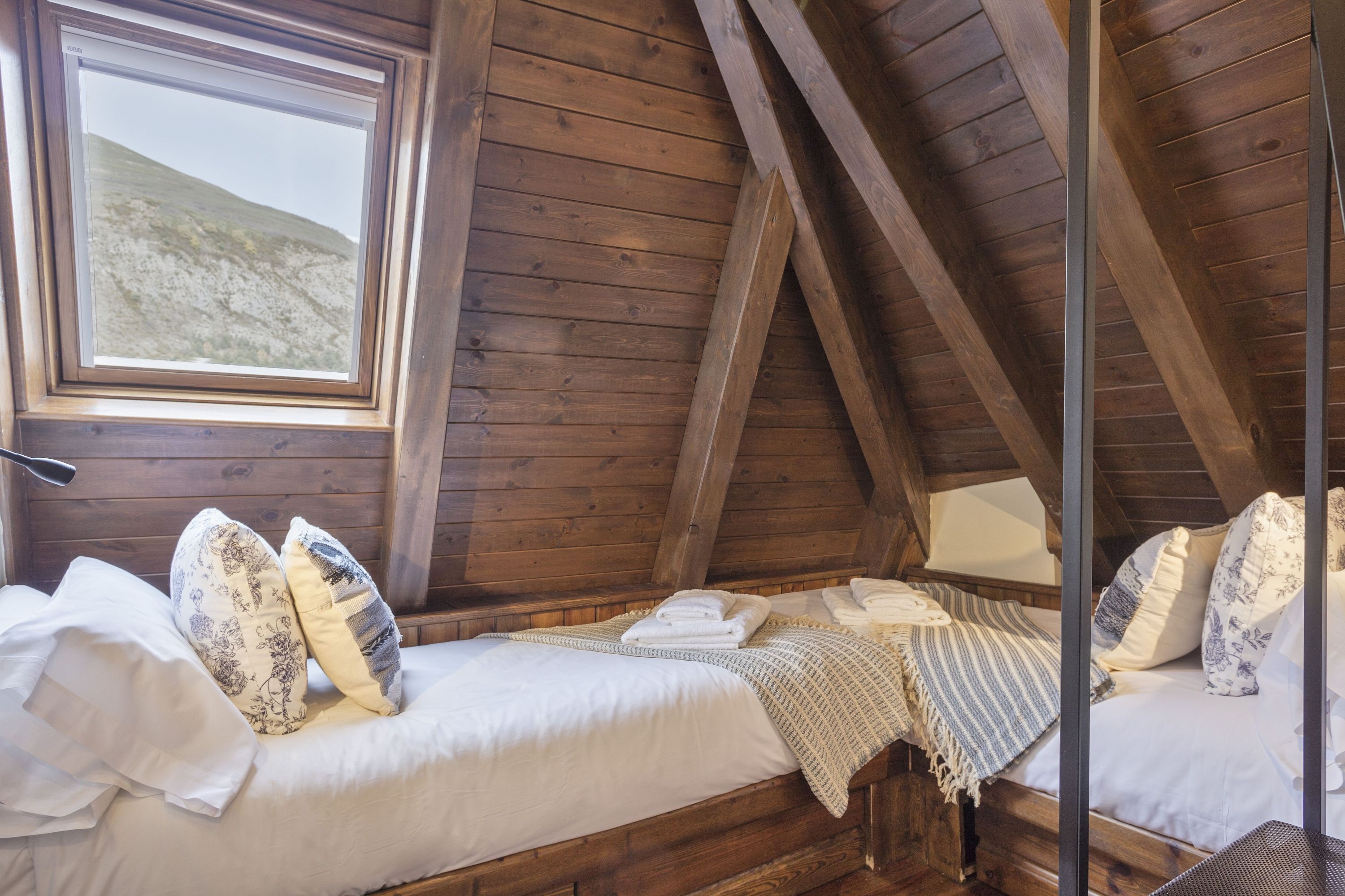 A cozy attic bedroom with wooden beams, a large window, and a bed with white linens and patterned pillows.