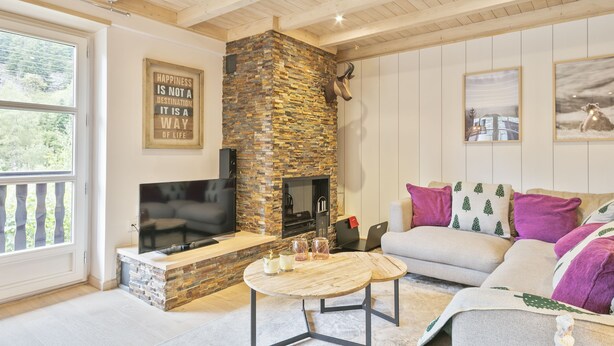 A cozy living room with a stone fireplace, a flat-screen TV, a sofa with decorative pillows, and a large window. - Luderna - Val de Ruda B52 dera Montjòia (Baqueira)