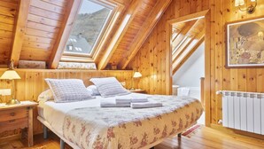 A bedroom with a wooden ceiling, a bed with a floral bedspread, two bedside lamps, and a painting on the wall. - Luderna - Dúplex Gessa C9 dera Montanheta (Gessa)
