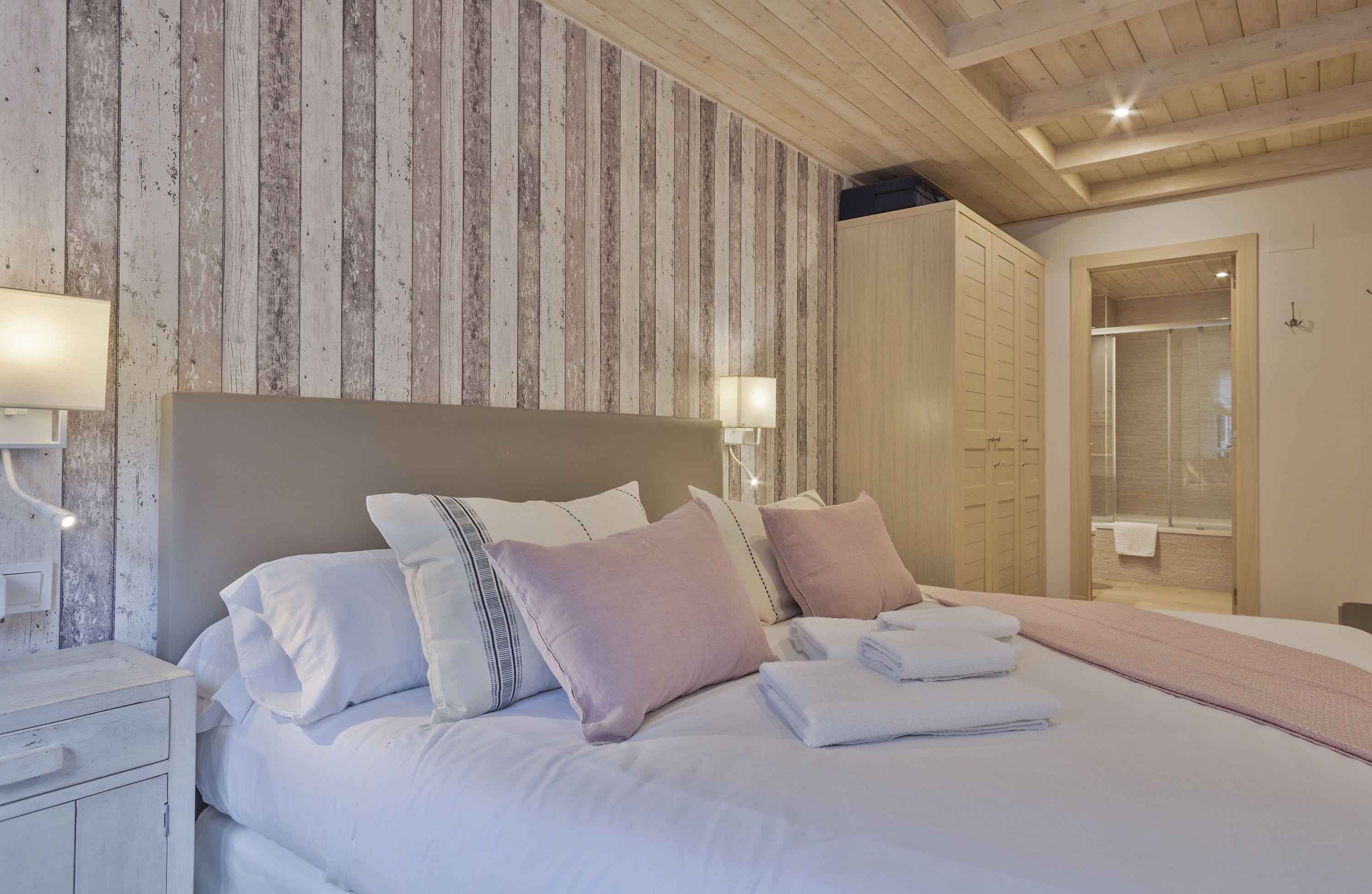 A neatly made bed with pillows and folded towels, a bedside lamp, and a wooden ceiling.