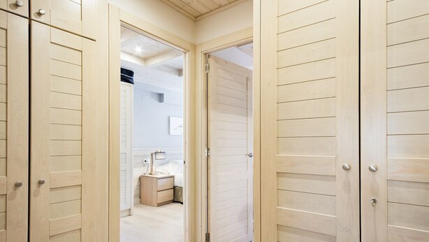 Un couloir avec des portes en bois menant à une chambre à coucher équipée d’un lit, d’une table de chevet et d’une lampe.