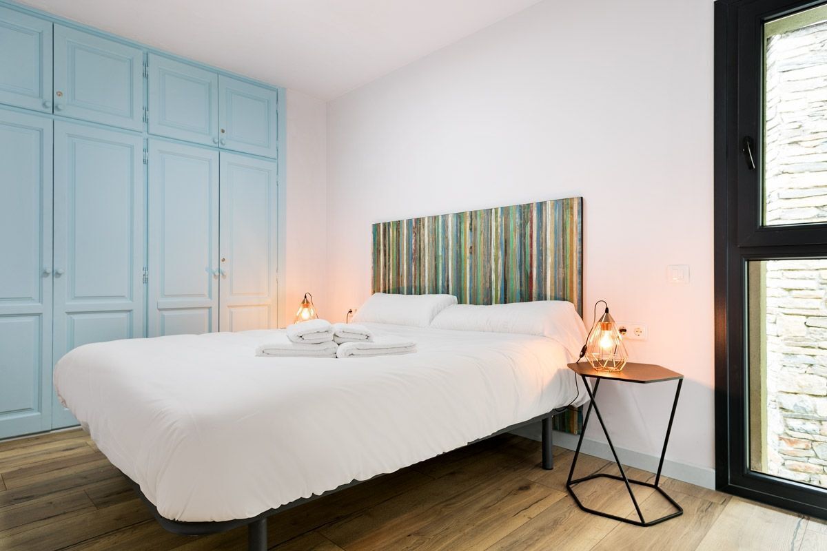 A bedroom with a large bed, a headboard with a striped pattern, two bedside lamps, and a window with a stone wall view.