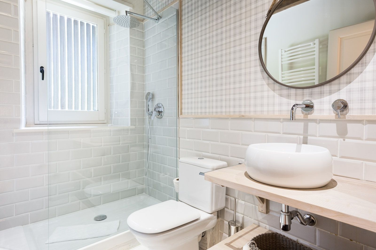 Een moderne badkamer met een glazen douchecabine, toilet, wastafel met spiegel en een raam met zonwering.