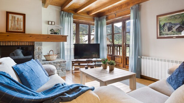 Una sala de estar acogedora con chimenea, vigas de madera, televisor y ventanas amplias con cortinas.