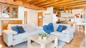 Un salón acogedor con un sofá de color gris claro, vigas de madera, una pequeña mesa de centro de madera con plantas y una cocina al fondo.