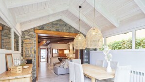 A spacious dining area with a wooden table and white upholstered chairs, a stone wall, and a hanging rattan light fixture. - Luderna - Casa con jardín Vista Aneto (La Val d'Aran)
